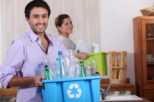 A young man with dark hair, wearing a light purple shirt, is smiling and holding a blue recycling bin filled with empty glass bottles and jars of various shapes and green, clear, and brown tones, situated in a domestic kitchen environment. The background features a woman with dark hair in a loose bun, dressed in a light-colored top, standing near a green plastic container for waste sorting, with wooden kitchen cabinets, a wicker basket, and a wooden shelving unit visible behind her. The scene is well-lit with natural and indoor lighting, emphasizing the textures of the glass bottles, the smooth surface of the bins, and the warm tones of the wooden furniture, reflecting a typical household setting focused on rubbish sorting and recycling. The image is associated with domestic rubbish disposal and recycling services, such as those provided by Havering House Clearance, in the local area.