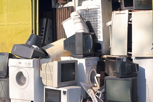 A collection of discarded household appliances and electronic waste piled together outdoors on a concrete surface, including washing machines, computer monitors, and microwave ovens, with some items stacked or leaning against each other. The appliances differ in size, material, and color, predominantly in shades of white, black, and beige, with visible textures such as plastic casings, glass screens, and metal panels. The background features a dark, industrial setting with metal storage units and a yellow wall, indicating an area used for rubbish collection or clearance. The environment suggests a waste management or clearance situation, consistent with services provided by Havering House Clearance, with natural daylight illuminating the scene and highlighting the clutter of junk intended for disposal or recycling in a suburb nearby London.